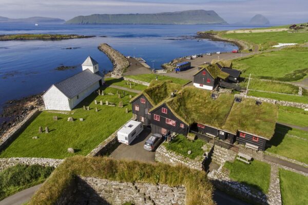 Scenic village by the sea with grass-roofed houses and a white church, surrounded by lush green fields and mountains.