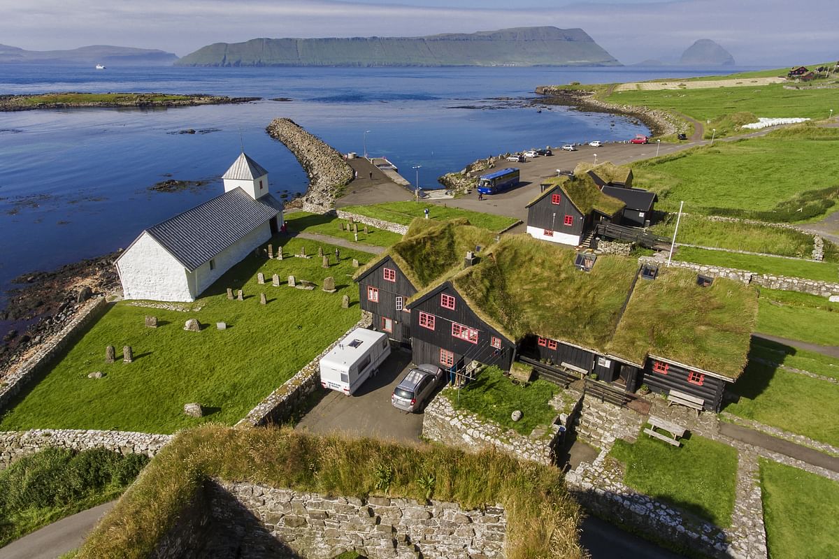 Scenic village by the sea with grass-roofed houses and a white church, surrounded by lush green fields and mountains.