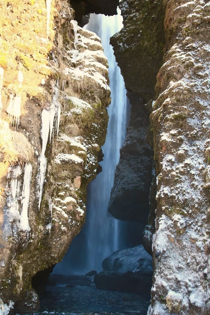 Narrow canyon waterfall with icy cliffs and winter vegetation, sunlight highlighting the rocky passage.
