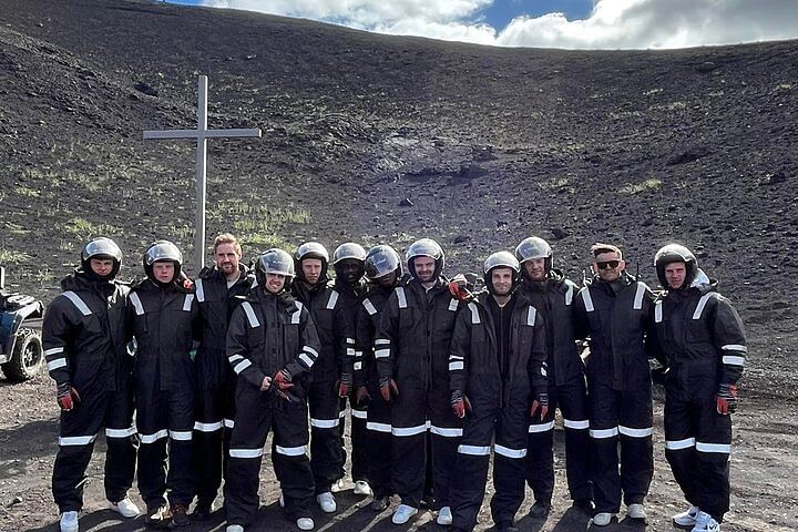 Group in protective gear standing near a cross on a rocky landscape under a cloudy sky.