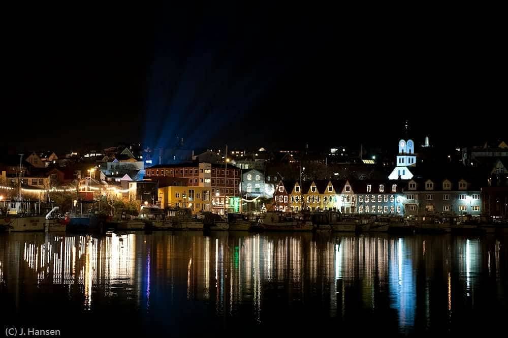 Nighttime harbor with illuminated buildings and reflections on water, highlighting a picturesque coastal village.