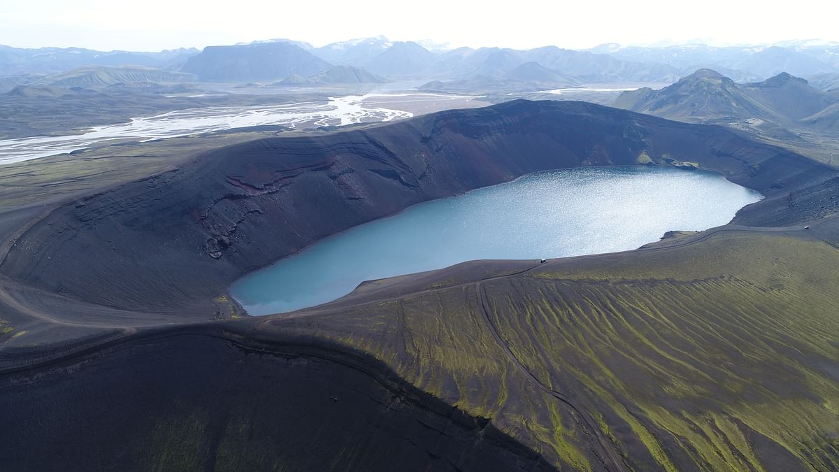 Aerial view of a volcanic crater lake surrounded by rugged terrain in a remote mountainous landscape.