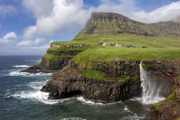 Scenic coastal view of rocky cliffs, waterfall, and grassy hills with scattered houses under a blue sky with clouds.