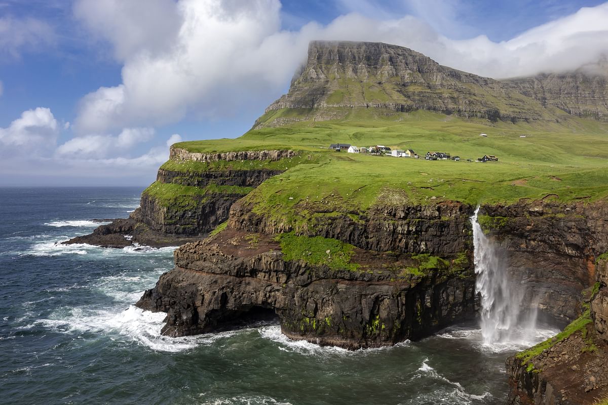 Scenic coastal view of rocky cliffs, waterfall, and grassy hills with scattered houses under a blue sky with clouds.