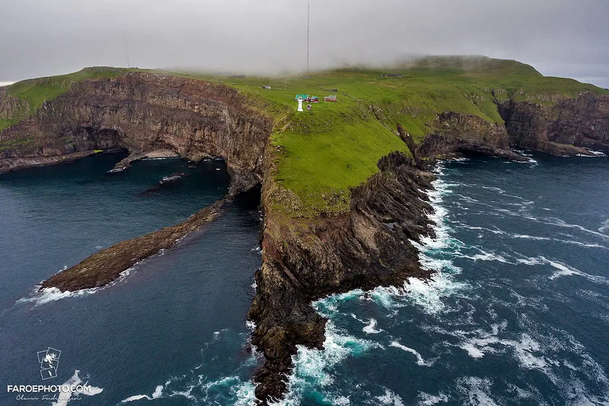 Aerial view of rocky cliffs and lush green landscape meeting ocean waves under a misty sky in the Faroe Islands.