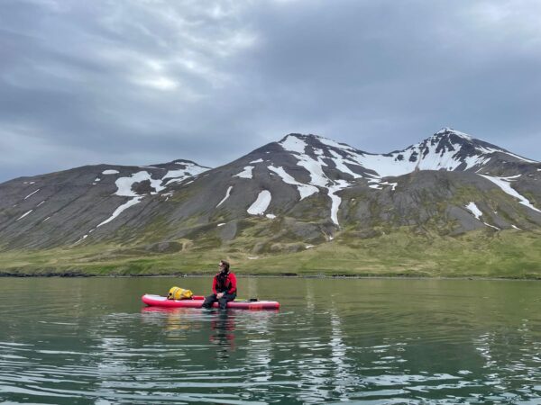 Person paddleboarding on calm water with snow-capped mountains in the background under a cloudy sky.