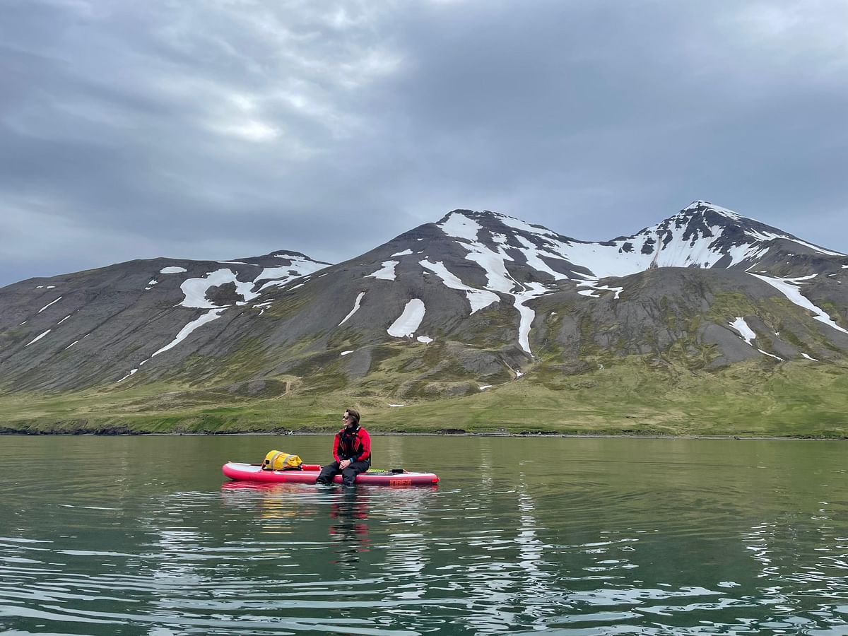 Person paddleboarding on calm water with snow-capped mountains in the background under a cloudy sky.