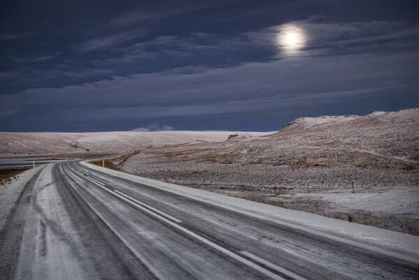 Desolate snowy road under a cloudy night sky with visible moon, creating an eerie winter landscape.