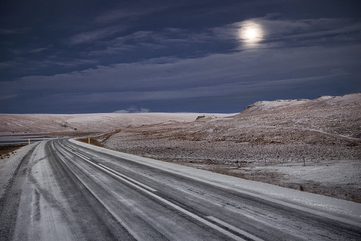 Desolate snowy road under a cloudy night sky with visible moon, creating an eerie winter landscape.