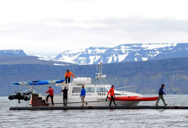 People boarding a fishing boat with kayaks, snowy mountains in the backdrop.