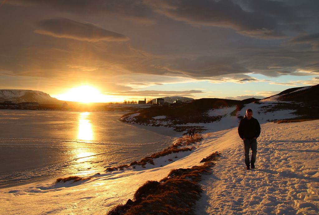 Person walking on a snowy path by a frozen lake at sunset, with dramatic clouds and reflections on the water.