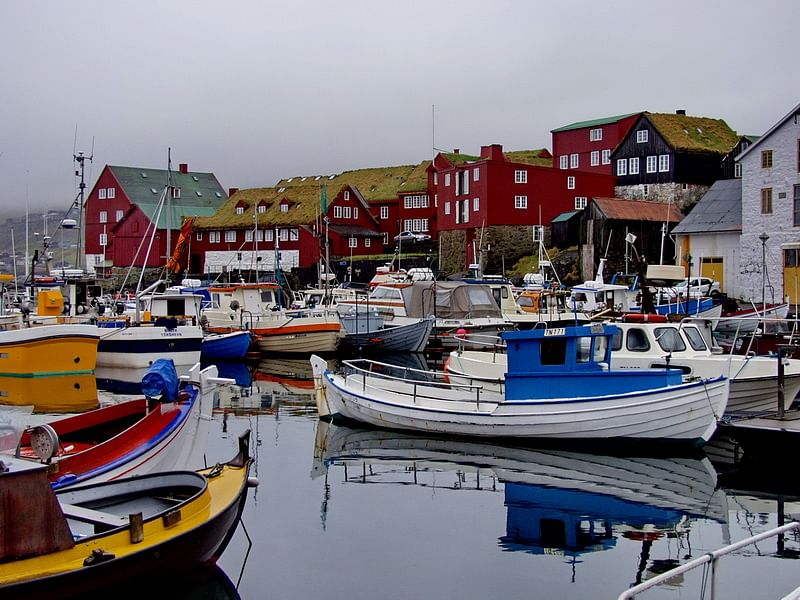 Colorful boats docked in a marina with traditional grass-roofed red buildings in the background on a cloudy day.