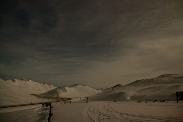 Snow-covered mountain landscape at night with a winding road and cloudy sky.