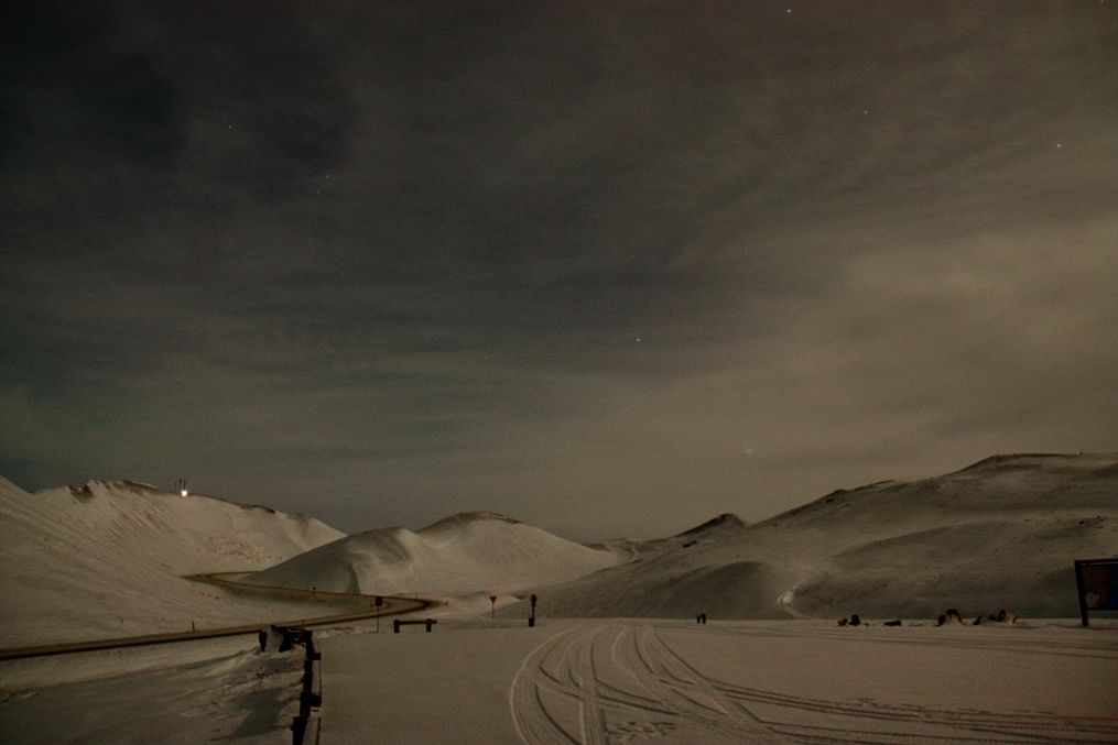 Snow-covered mountain landscape at night with a winding road and cloudy sky.