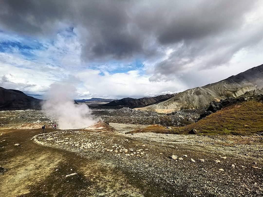 Rugged landscape with steam rising from geothermal vents, cloudy sky, and distant mountains in a volcanic terrain.