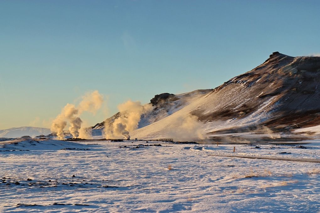 Snowy geothermal landscape with steaming fumaroles and mountains under a clear blue sky.