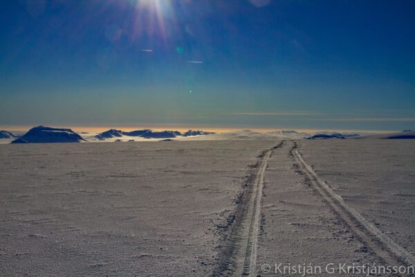 Snowy landscape with tire tracks leading to distant mountains under a bright sunlit sky.