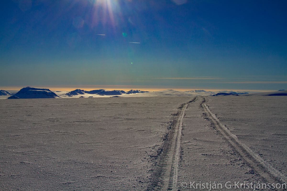 Snowy landscape with tire tracks leading to distant mountains under a bright sunlit sky.