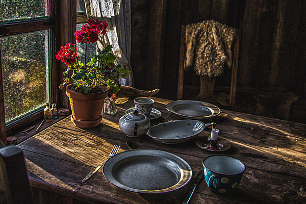 Rustic dining table with vintage crockery, potted geraniums, and sunlight streaming through a window. Cozy ambiance.