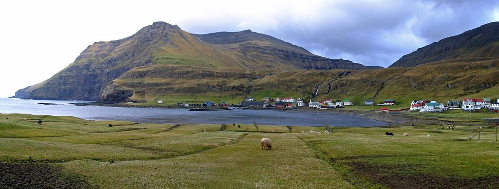 Scenic view of a grassy landscape with mountains and a small coastal village under a cloudy sky in the Faroe Islands.