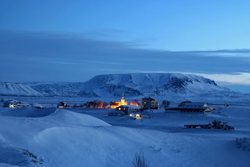 Snowy Iceland village at dusk, warm lights glowing, with mountains in the backdrop and a serene winter atmosphere.