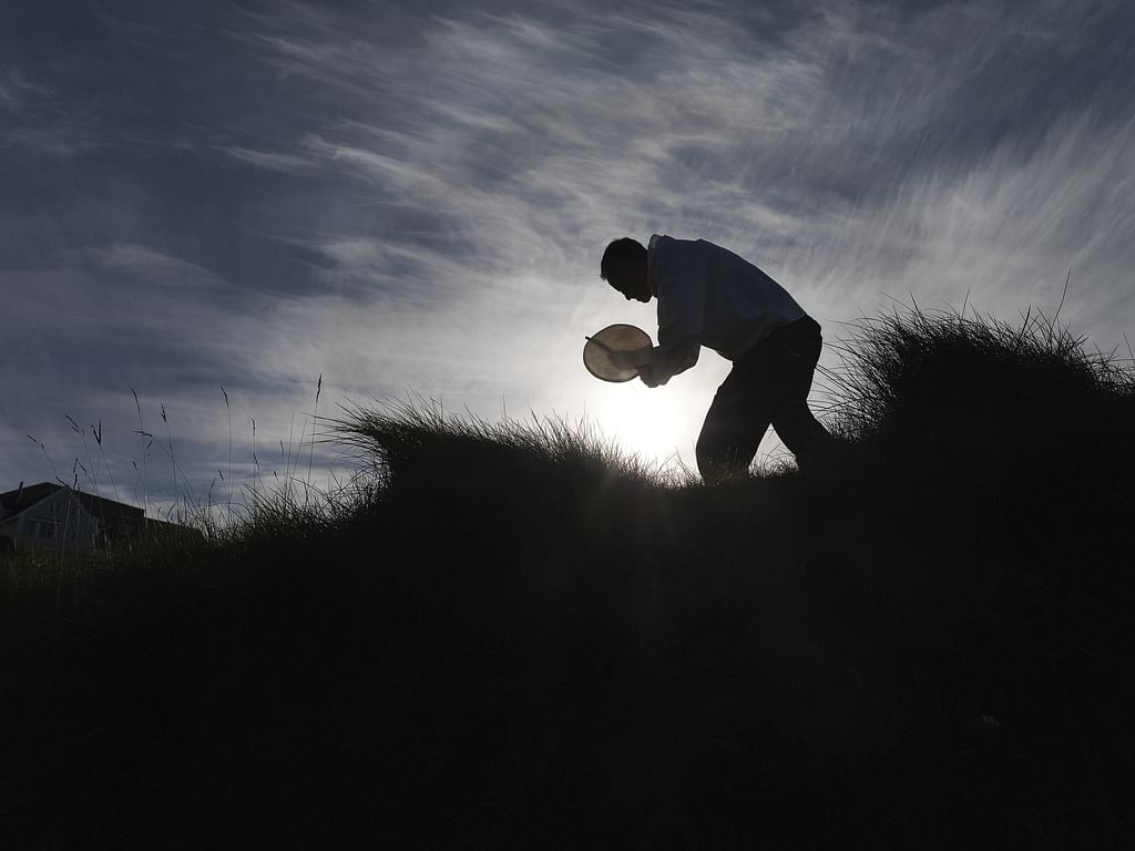 Silhouetted person playing tambourine on grassy hill at sunset.