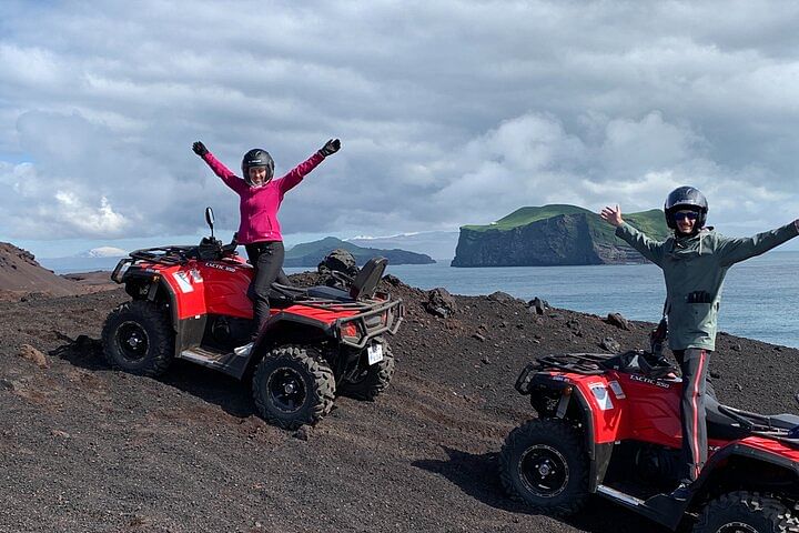Two people on ATVs celebrating on a rocky coastal landscape with an island in the background under a cloudy sky.