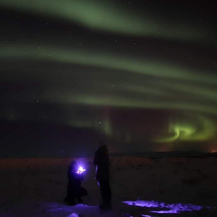 Northern lights illuminate the night sky with a couple, one kneeling, under the aurora's vibrant display.