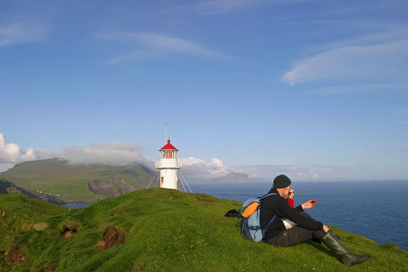 Hiker resting near Kallur Lighthouse on green cliffs, overlooking ocean and clear sky, Faroe Islands scenic view.