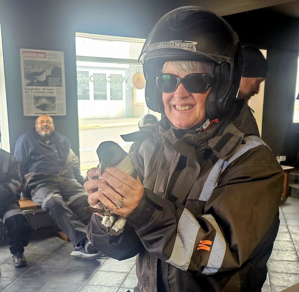 Smiling woman in motorcycle gear holding a small bird indoors, with people seated in the background.