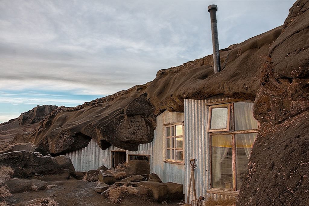 Rustic cliffside house with tin walls and chimney blends into rugged landscape under cloudy sky.