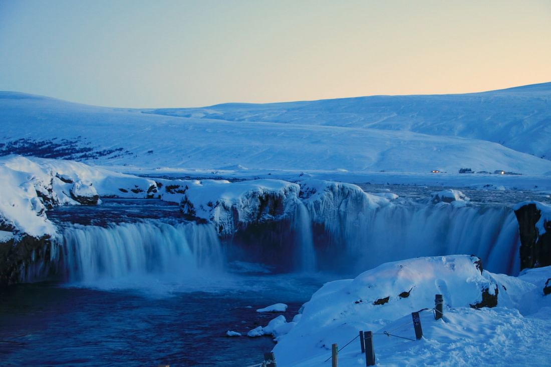 Snowy waterfall in Iceland during winter twilight, surrounded by icy landscape and distant hills.