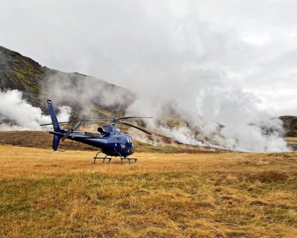 Blue helicopter landing on grassy field with steaming geothermal vents in the background.